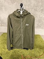 Front view of olive green hooded jacket with Dior logo on left chest, displayed on hanger against concrete wall and green rug.