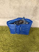 Blue bag filled with jeans and pants grouped inside, on green rug against a concrete wall.