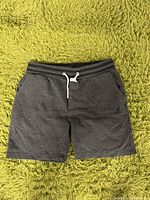 Front view of grey Kangol men's sweat shorts laid flat on green fluffy surface, showing front pockets, drawstring waistband, and Kangol logo.