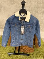 Front view of kids denim jacket showing sherpa collar, two chest flap pockets with buttons, and front button closures, hanging on black hanger with green carpet background.