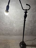 Tall black metal floor lamp without shade with light bulb attached, shown standing against a grey wall.