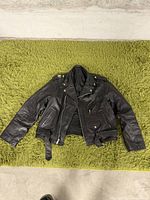 Front view of black leather motorcycle jacket with zippered pockets, snap buttons on collar, and belt at waist, laid flat on green carpet.