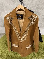 Front side of brown suede jacket with white embroidered floral pattern on chest and pockets, two-button closure, hanging on wooden hanger.