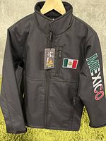 Front view of men's black softshell jacket with Mexico flag patch on chest, zippered pocket, and MEXICO text on right sleeve.