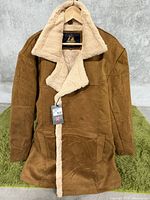 Front view of men's tan suede long coat with fur collar and lining on green fuzzy surface, showing double-breasted buttons and front pockets.