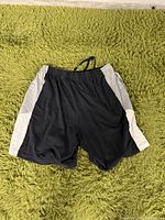 Front view of black shorts with gray side panels on sides lying on green carpet.
