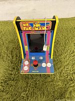 Front view of Ms. Pac-Man mini arcade game showing joystick, buttons, and screen area with colorful artwork and logo.