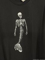Front view of black T-shirt showing white skeleton mermaid graphic centered on front.
