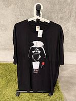 Full view of black Darth Vader T-shirt hanging on hanger against concrete wall and green carpet.