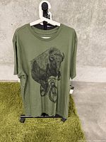 Full view of olive green T-shirt hanging on a hanger against a concrete wall and greenish carpet