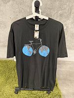 Front view of black T-shirt hanging on white hanger showing bicycle graphic with world map styled wheels.