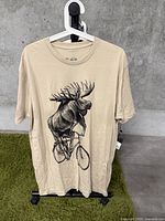 Front view of beige t-shirt hung on hanger against concrete background showing moose riding bicycle graphic.