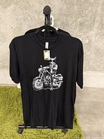 Front of black t-shirt on hanger against wall and green carpet, showing graphic of cat on motorcycle.
