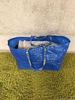 Blue IKEA bag filled with folded pants and jeans on a green rug against a grey wall.