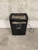 Black paper shredder on floor, view from front, showing power cord and device shape.