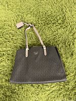 Front view of the dark brown Guess tote bag with beige handles, lying on a green textured surface.