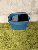 Blue plastic bin filled with folded clothing items, visible top items include a folded gray sweatshirt and a black garment, placed on a green carpet against a concrete wall.