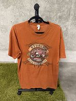 Front view of orange T-shirt with Sturgis 2012 Black Hills Rally motorcycle graphic on hanger.