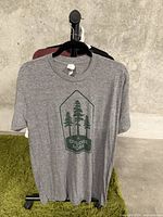 Full view of grey T-shirt hanging on hanger displaying the front graphic with trees and text