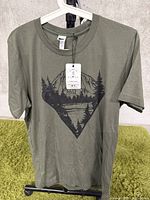 Front view of a green T-shirt with a printed black nature-themed graphic showing trees, mountains, and water inside a diamond shape. The T-shirt is on a white hanger against a concrete background.