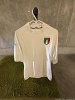 White Italia T-shirt hanging on a hanger in a concrete wall background.