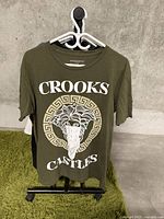 Front view of an olive green Crooks & Castles T-shirt. Features a large white Medusa head graphic inside a circular Greek key border with the Crooks & Castles brand name above and below the graphic.