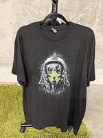Full view of dark gray XXL T-shirt displayed on hanger against a concrete background and green carpet.