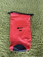 Red waterproof dry bag with black bottom and rolled top, lying flat on green carpet. 'Mountain Equipment Co-op' logo and '10 Litres' printed in black on front.