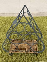 Photo showing triangular metal wine rack with circular holders and wicker base on green carpet outdoors against a wall.