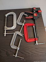 Four C-clamps of different sizes and colors arranged on dark wood surface.