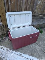 Red Coleman cooler with white lid, shown open to display interior, placed on concrete near wooden fence.