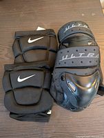 Photo showing two knee pads on a wooden surface: a black Nike foam padded knee pad and a black mesh knee pad with plastic cap and adjustable straps.