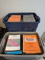 View of two plastic bins containing assorted vintage Toyota manuals including orange and gray softcover booklets.