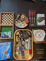 Full view of entire lot items on a dark surface including wooden chess/checkers board, fishing accessories, dart set, ear plugs, and outdoor thermometer with black box.