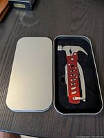 Photo of an open metal case containing a red multi tool with hammer head and other foldable tools inside on a wooden surface.