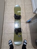 Full view of olive green Rome Anthem snowboard with black bindings, viewed from above on tiled floor, showing length and condition