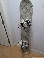 Full view of the Salomon snowboard standing upright showing black and white geometric grid design and attached white bindings.