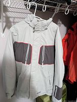 Full front view of the ski jacket showing light gray body with dark gray and red accent chest pockets and cuffs, hanging on a clothing rack.