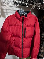 Full view of red Tommy Hilfiger puffer jacket on hanger showing front zipper and pocket with logo.