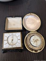 Two travel clocks pictured side by side on a dark surface, one square with gold-tone markers and the other round with world time zone dial and map, both with their cases open.