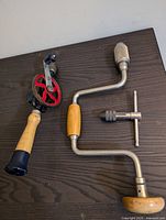 Two vintage hand drills on a dark surface, one red gear-driven and one brace drill with chuck key.