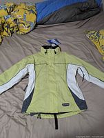 Light green waterproof jacket with white and dark gray panels spread flat showing front side and collar with tags attached.