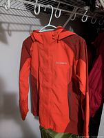 Front view of orange Columbia woman's rain jacket with hood hanging on white hanger showing color and design.