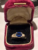 Ring displayed in red velvet jewelry box, showing square blue stone and engraved shoulders