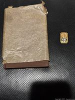 Photo showing 1927 book with worn protective paper jacket and rectangular Masonic pin with letter 'M' on a black surface.