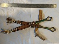 Two sharp metal tools with greenish handles crossed on white background with leather sheaths underneath, a metal ruler placed above for scale.