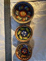 Three small decorative bowls with intricate beadwork in geometric and floral patterns, shown side by side with a ruler for size reference