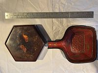 Two lacquer pieces side by side with a ruler for size reference. The hexagonal box shows a dark reddish-brown color with gold designs.