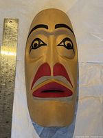 Front view of the carved wooden First Nations mask next to a metal ruler showing size approximately 9 inches tall. Mask features red mouth and black painted eye and eyebrow details.