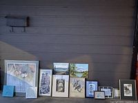Group photo overview of all 13 pieces in the lot, arranged against a wall, showing a mixture of framed artwork and a single blue decorative tile or ceramic piece.